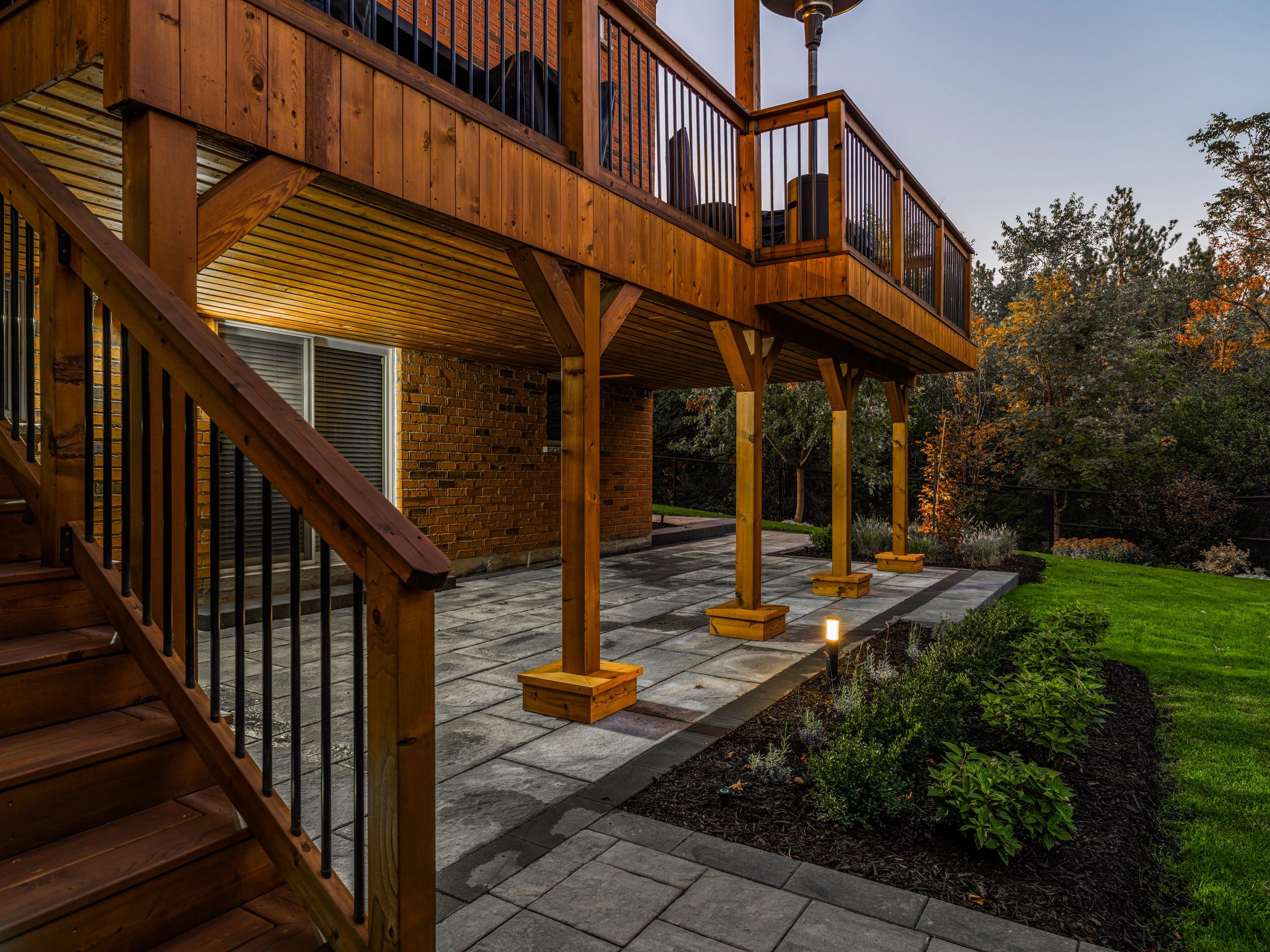 Wooden deck with stairs overlooking a paved patio and garden at dusk.