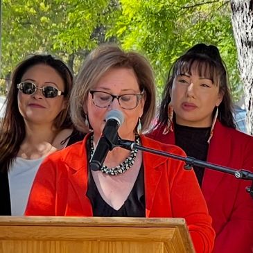 A woman speaks at a podium with officials standing behind her outdoors.