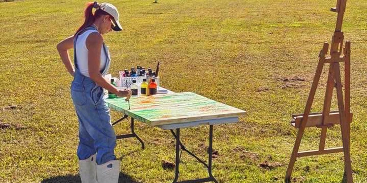 Woman painting outdoors on a sunny day, surrounded by grass.