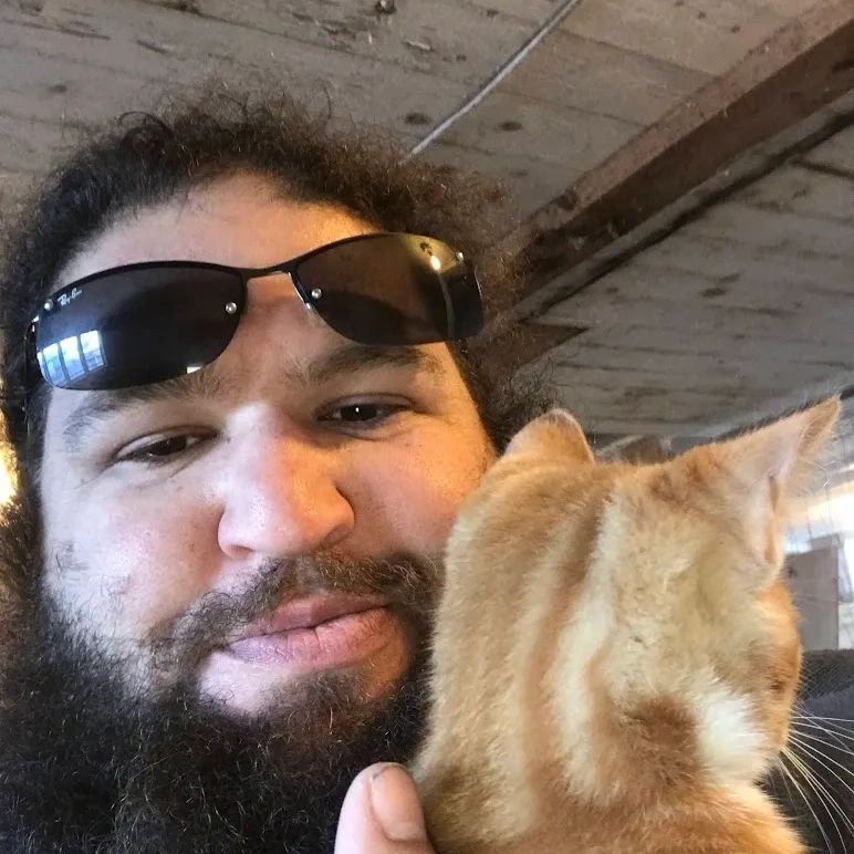 Image shows Farmer holding an orange cat close to his face, sunglasses on his forehead.