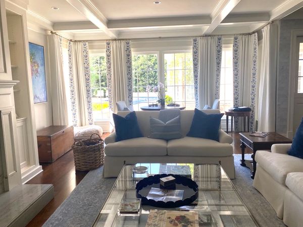 Bright living room with white couches, blue pillows, and large windows with floral curtains.