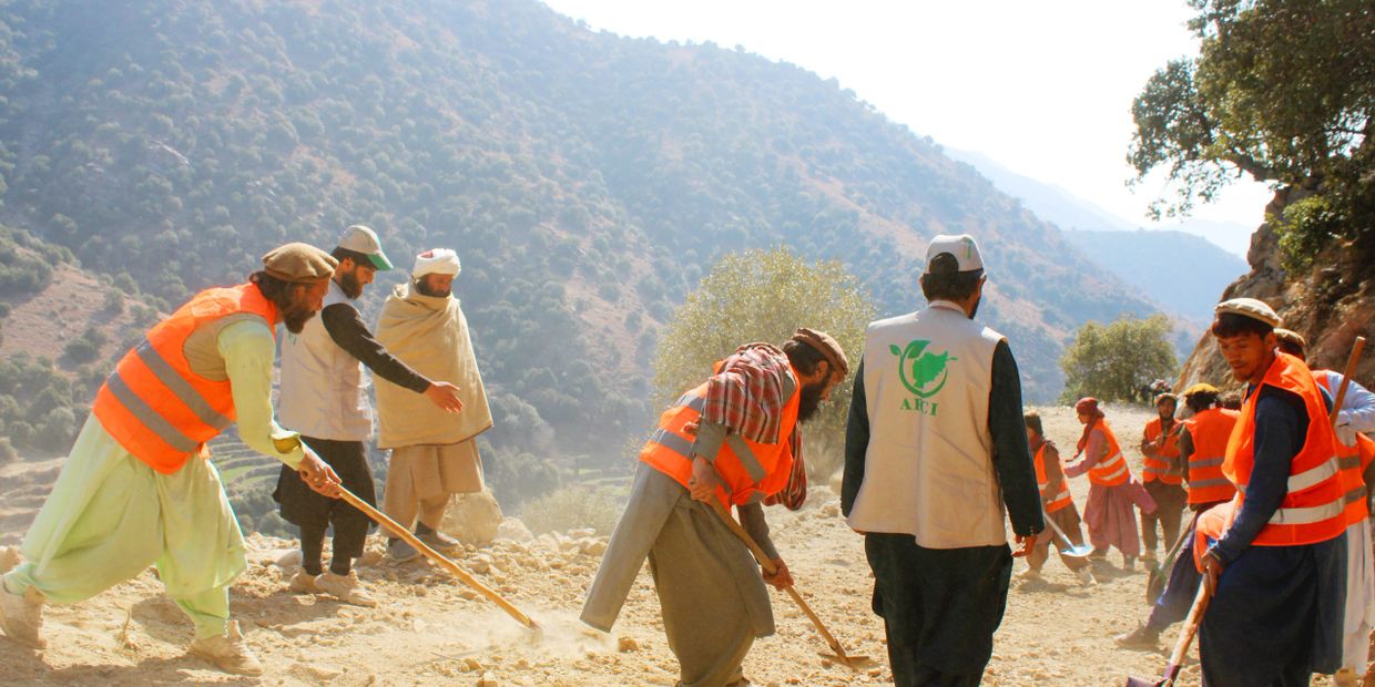 Group of men working on a rocky path in a mountainous area wearing orange safety vests.