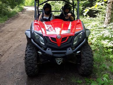 Two people riding a red off-road vehicle on a dirt trail surrounded by trees.