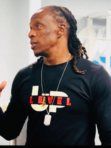Man with braided hair wearing a black shirt with "UP LEVEL" text and dog tags.