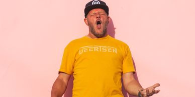 Man in yellow shirt holding skateboard and making an expressive face against a pink background.
