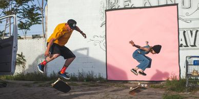 Two people performing skateboard tricks in front of a mural with a pink square.