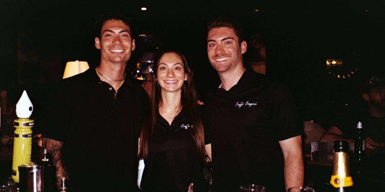 Three smiling bartenders in black shirts posing behind a bar with drinks.
