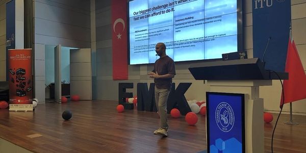 A man presenting on stage in a modern conference room with a large screen and Turkish flags.