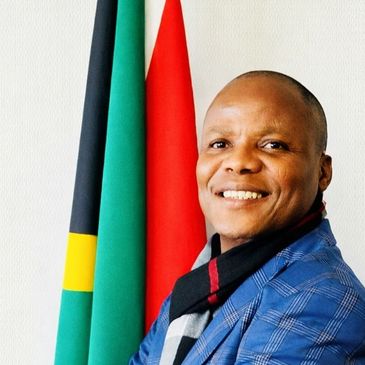 Smiling man in a blue checkered jacket standing beside two flags.