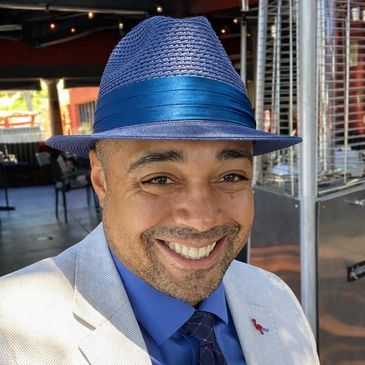 Smiling man wearing a blue hat and light-colored suit in an outdoor setting.