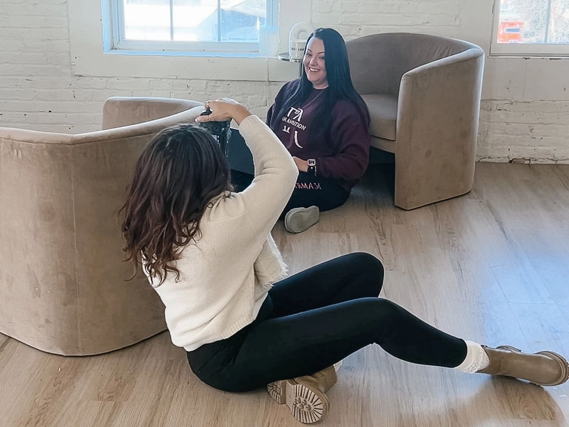 A photographer takes a picture of a smiling woman sitting on the floor indoors.