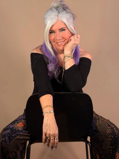 Smiling woman with purple-tinted hair sits backward on chair, wearing black off-shoulder top and patterned pants.