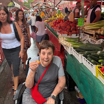 Elderly woman in wheelchair tasting fruit at a bustling outdoor market.