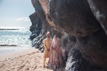 Couple in traditional attire on a beach by rocks.