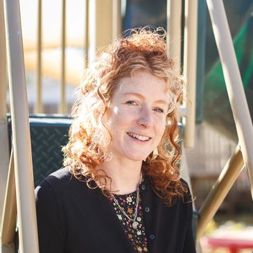 A smiling woman with curly red hair sitting on playground equipment in sunlight.