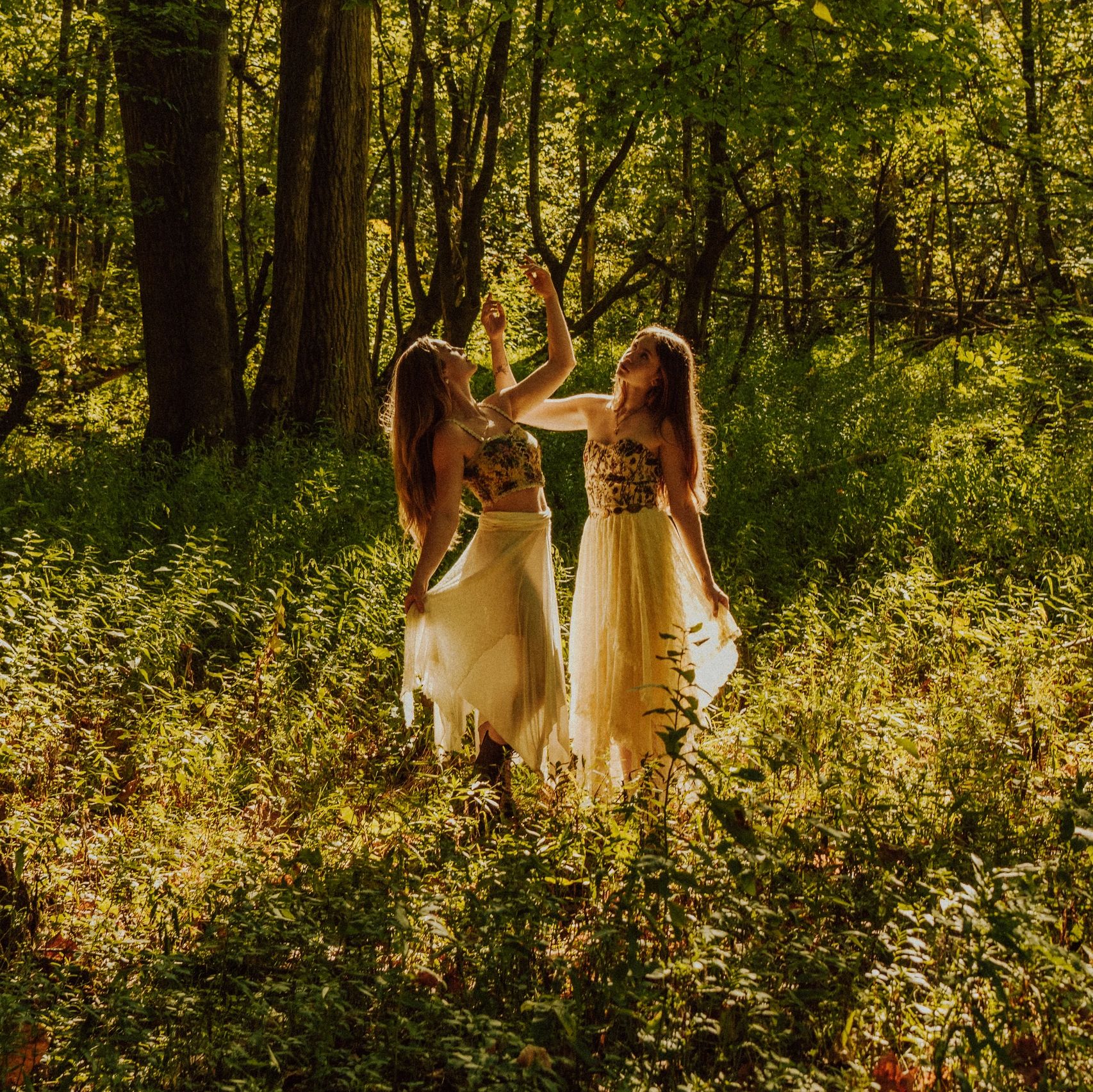 Two women in flowing dresses dance gracefully in a sunlit forest clearing.