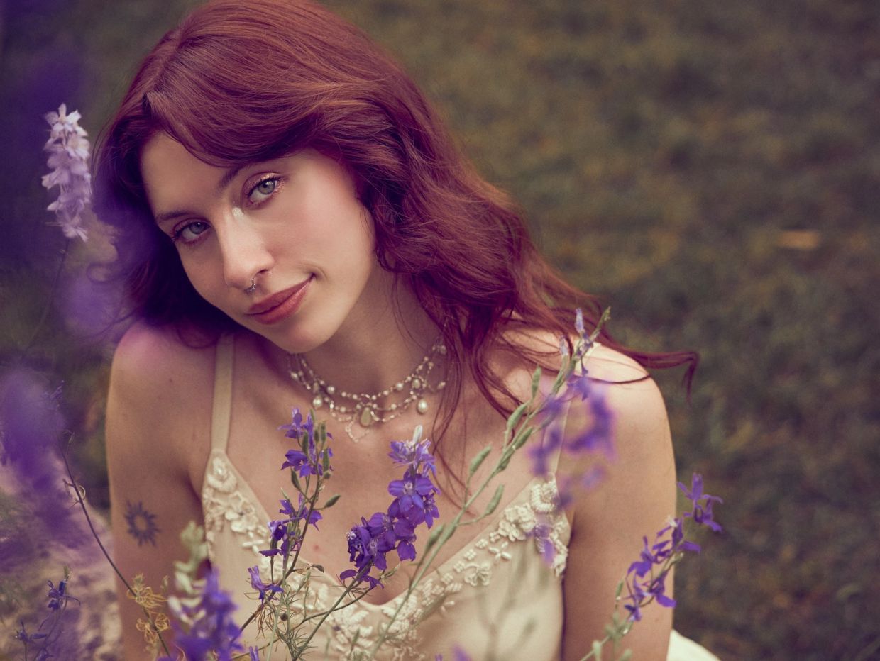 Red-haired woman in a beige dress with purple flowers around her.