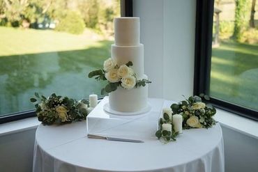 3 tier white wedding cake with greenery and white roses on table.