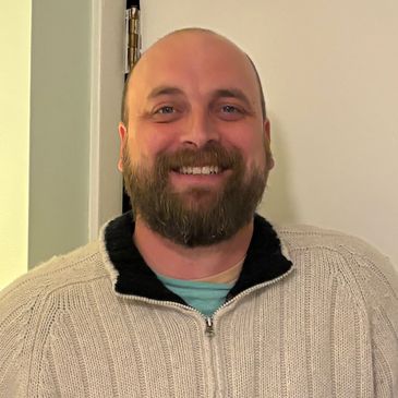 Smiling man in a beige knit sweater standing indoors.