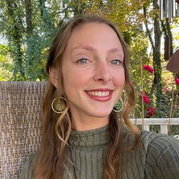 Young woman with wavy hair smiling outdoors with autumn foliage in the background.