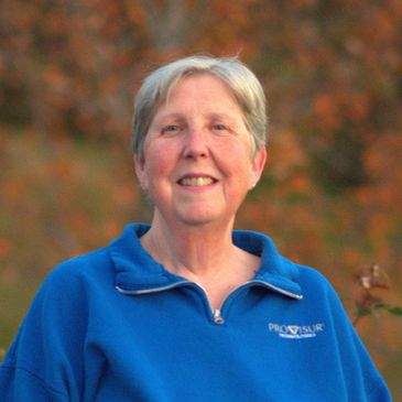 Smiling elderly woman in a blue sweatshirt outdoors with autumn foliage.