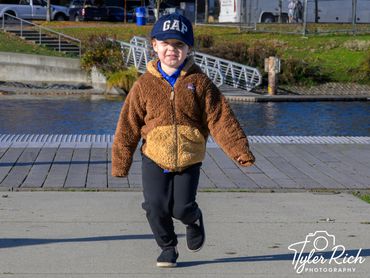 A young child in a brown jacket and GAP hat walking outdoors near water.