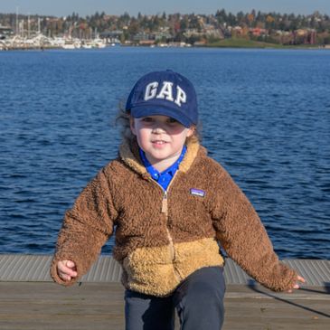 Young child in a brown jacket and blue GAP cap by the water.