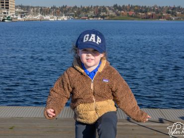 Young child in a brown jacket and blue GAP cap by the water.