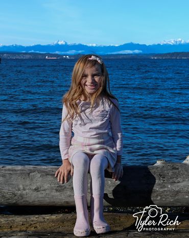 Smiling girl in pink boots sits on a log by the water with snowy mountains in the background.
