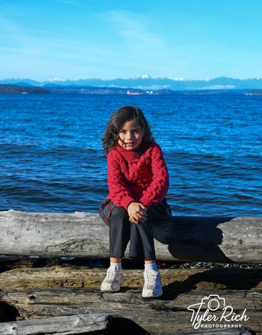 A young girl in a red sweater sits on a log by the blue sea with mountains in the background.