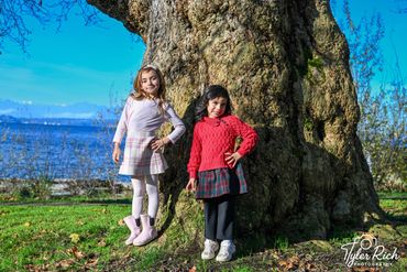 Two girls posing cheerfully by a large tree near a lake on a sunny day.