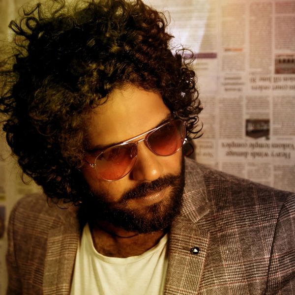 Man with curly hair and beard wearing sunglasses and a plaid blazer.