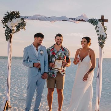 A joyful beach wedding with a bride, groom, and officiant under a decorated arch.