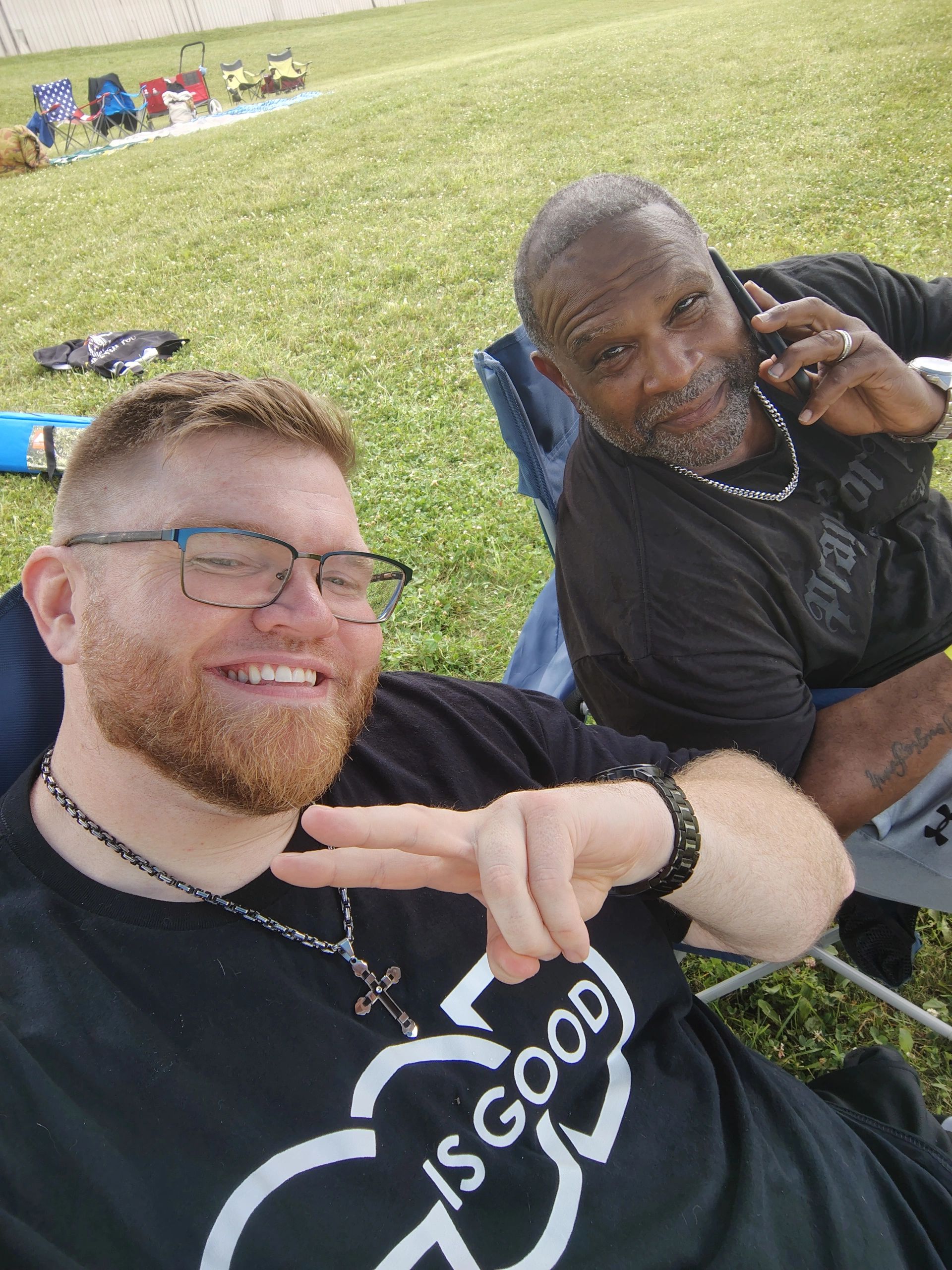 Two men sitting outdoors, one smiling and flashing a peace sign, the other on the phone.