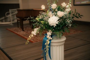 Elegant white floral arrangement with greenery in a white vase accented by a blue ribbon.