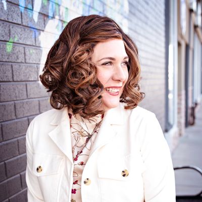 Smiling woman with curly hair wearing a white jacket and green pants.