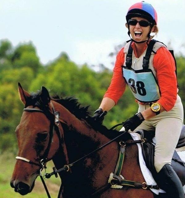 A smiling equestrian in gear riding a brown horse outdoors.