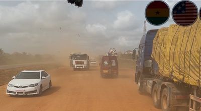 Vehicles drive on a dusty dirt road under a cloudy sky with Ghana and US flags.