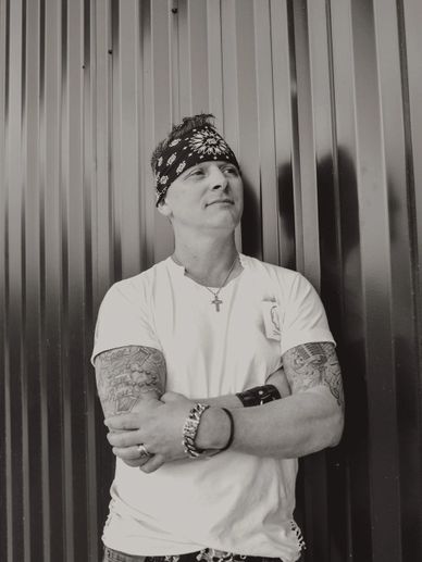 Man with tattoos and bandana stands confidently against a metal wall.