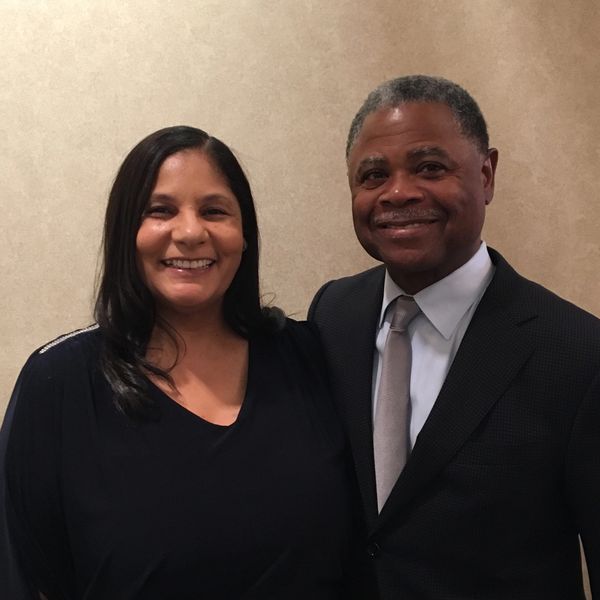 Two people smiling, dressed in formal attire against a beige background.