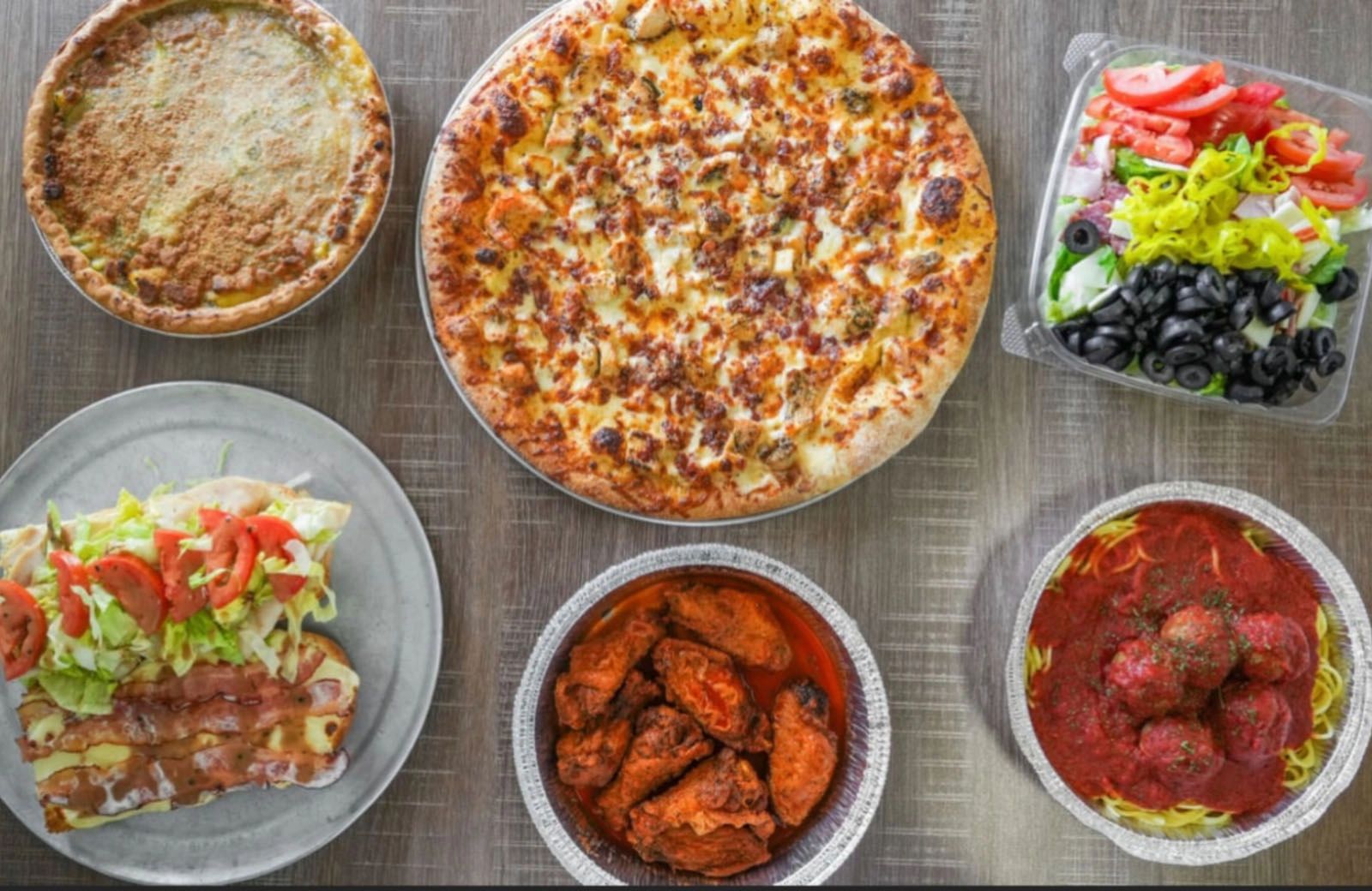 Picnic basket items on a table. Dessert pie, pizza, salad, sandwhich, wings, and pasta plate.