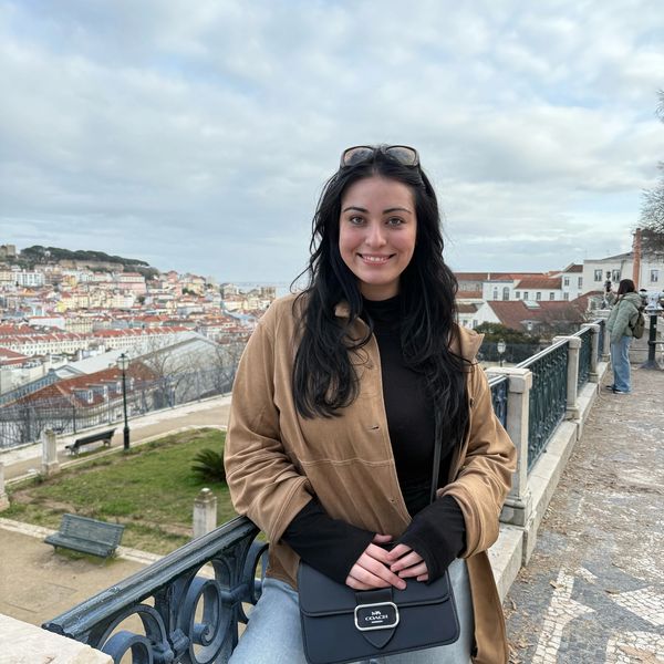 A woman in a brown jacket smiles near a scenic city viewpoint on a cloudy day.