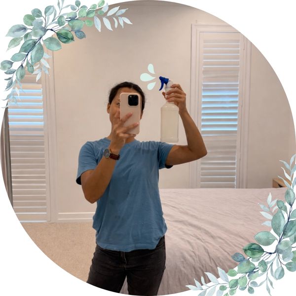 Person holding a spray bottle, taking a mirror selfie in a bedroom.