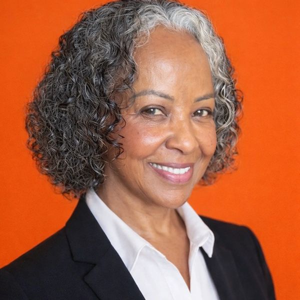 Confident older woman with curly salt-and-pepper hair smiles against an orange background.