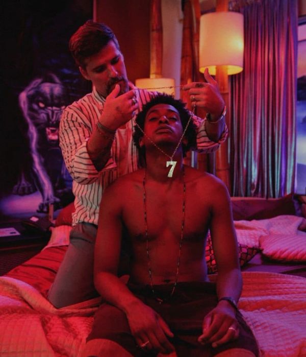 Man places a large '7' necklace on a shirtless man in a dimly lit room.