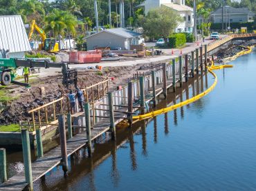 Construction work along a waterfront with pilings and safety barriers.