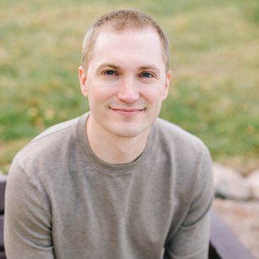 Young man with short hair smiling outdoors on a bench.