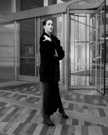 Woman in black coat posing in front of glass doors.