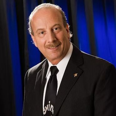 A smiling man in a black suit and tie against a blue backdrop.