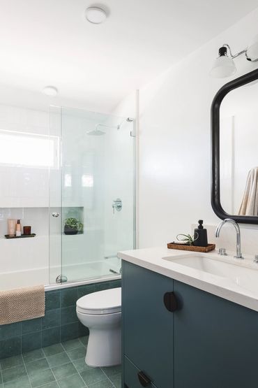 Modern bathroom with green floor tile, teal vanity, glass shower enclosure & white walls. 